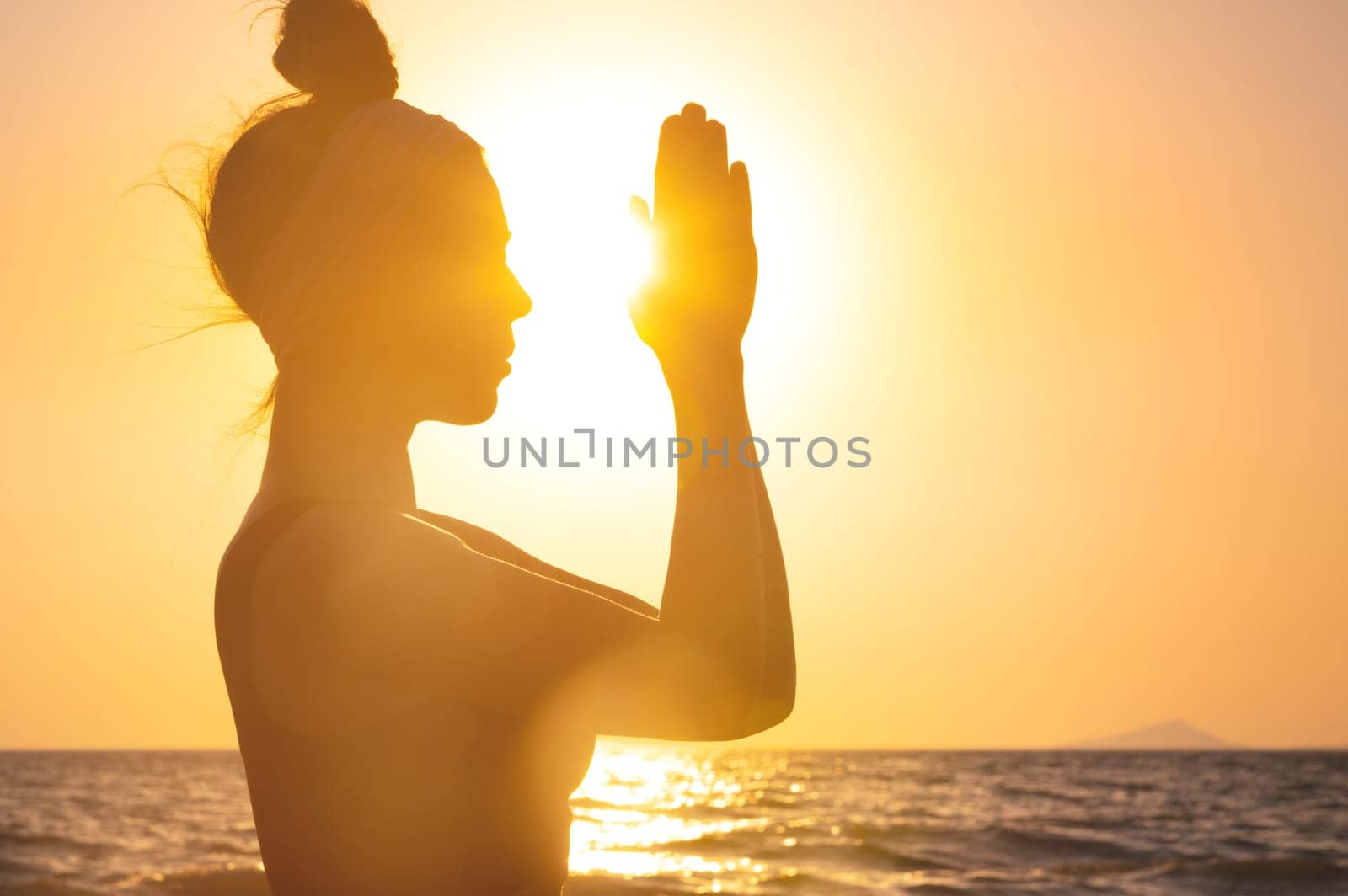 Silhouette of a woman sitting on the shore and praying with her hands folded. Woman with folded hands on the seashore during sunset or dawn practicing spiritual activities.