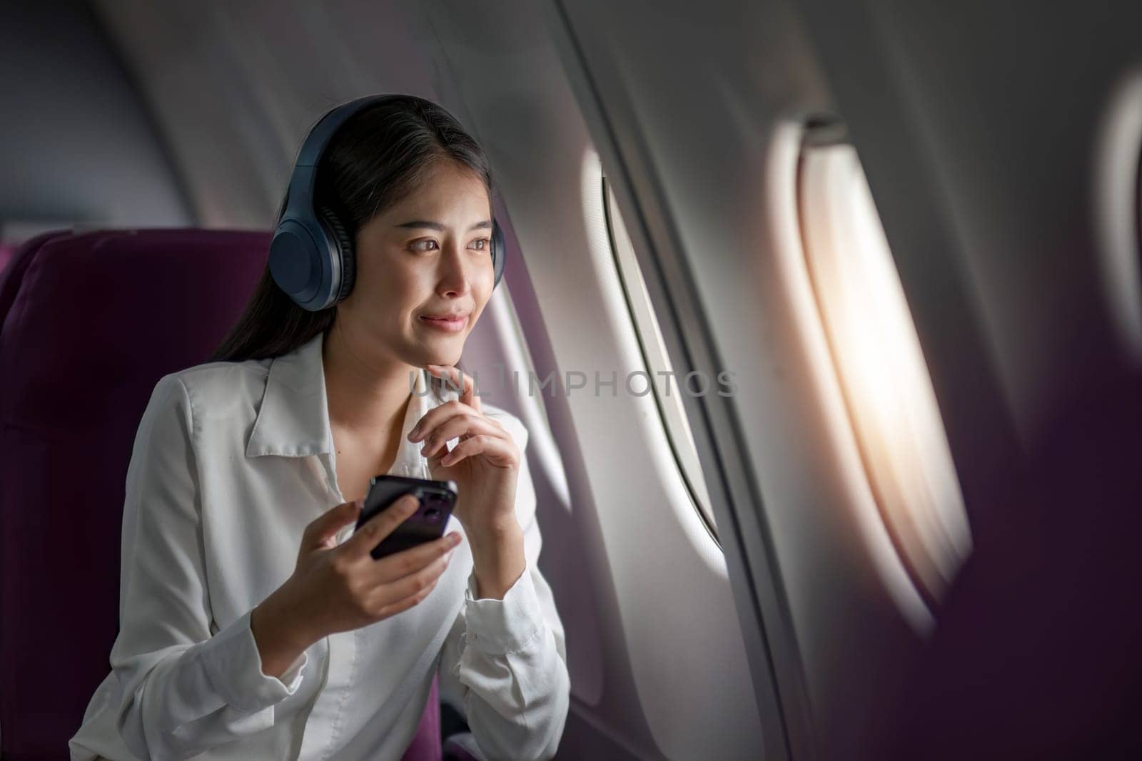 Asian woman sitting and using a smartphone in an airplane Listening to music next to the airplane seat window travel and technology concept.