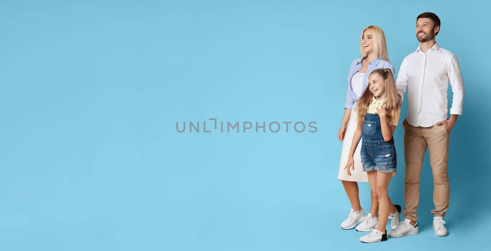 A happy family of three stands together in front of a light blue backdrop. The parents smile at the camera, while the young daughter looks off to copy space