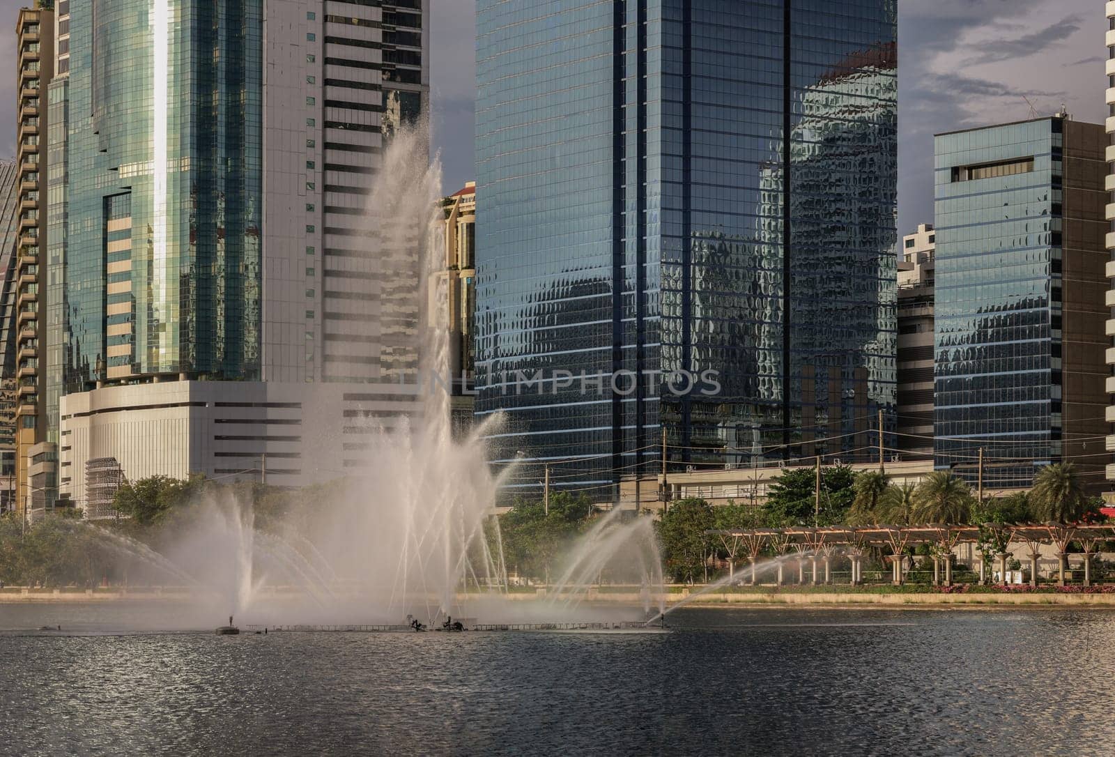 Bangkok, Thailand - 22 Jun, 2024 - Cityscape office buildings with modern architecture with pond water fountains. Picturesque view of skyscrapers and dramatic sky before sunset. Copy space, Selective focus.