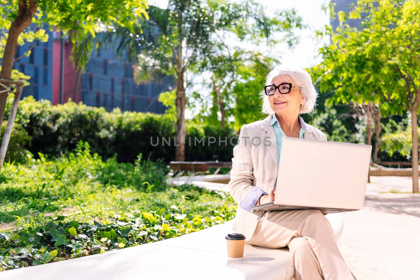 middle age woman smiling happy using laptop sitting outdoors in a city park, concept of technology and modern mature people lifestyle, copyspace for text