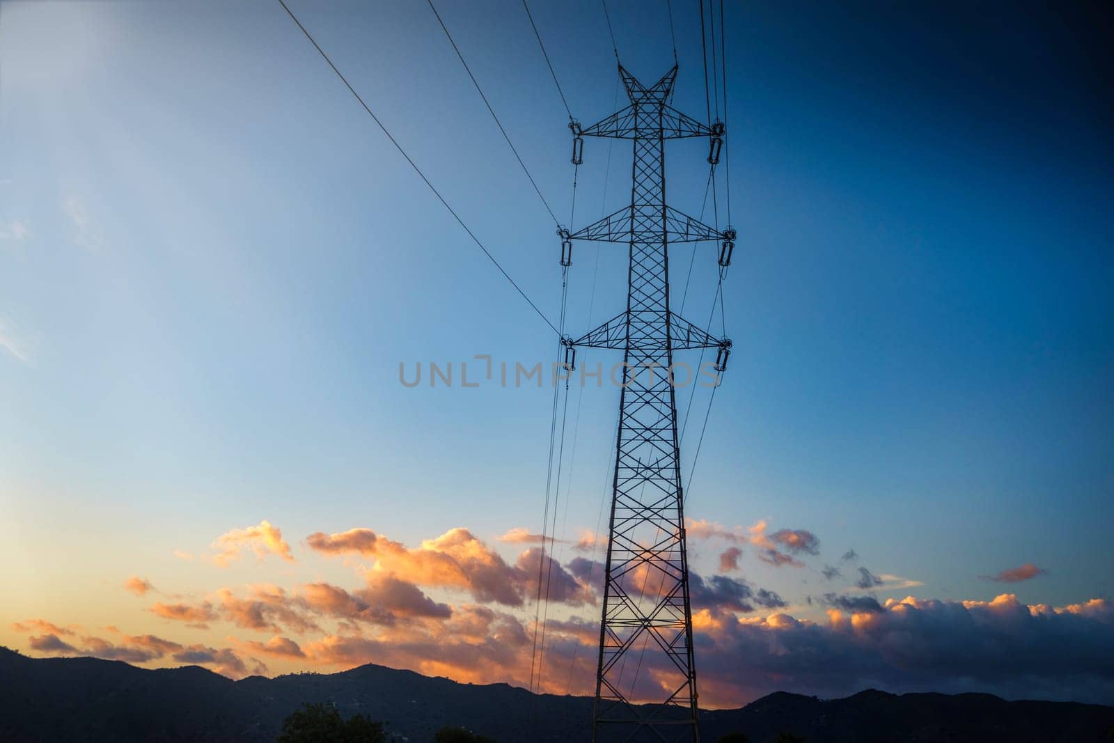 high-voltage electricity pylon (also known as a power tower or electric transmission tower) silhouetted against a sky exhibiting a beautiful palette of colors due to sunset or sunrise. Multiple power lines are connected to the pylon, extending out across the frame. In the background, a mountain range can be seen, adding layers to the horizon and further enhancing the scenic beauty of the photograph. The play of light and clouds in the sky creates a serene backdrop for the stark structure of the pylon.