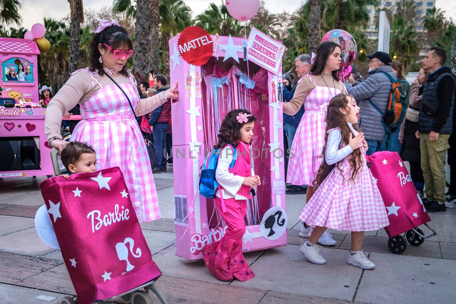 Barbie theme. There is a mix of adults and children all sporting variations of pink dresses or attire with the iconic Barbie logo. One adult is pushing a cart designed to look like a Barbie packaging box, and there are large bags with the Barbie logo being held or worn as backpacks as well