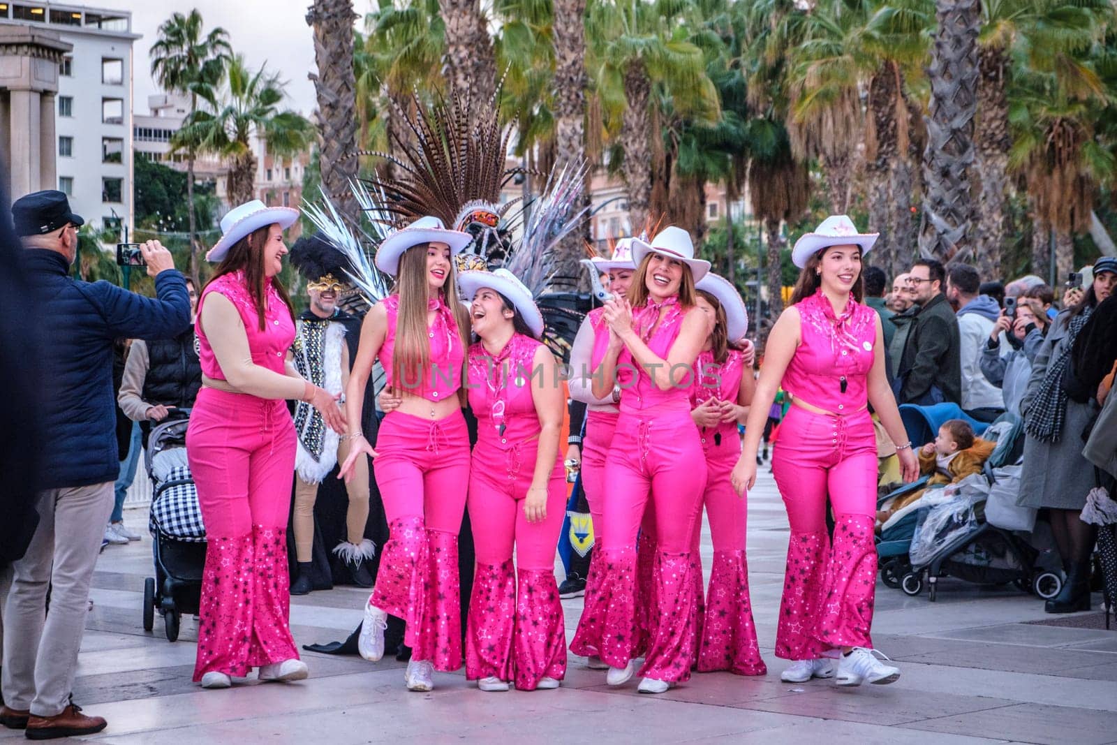 There are several people gathered in what looks like an outdoor public space with palm trees in the background. The focus of the image is on a group of people, predominantly women, who are dressed in matching bright pink outfits with white hats that resemble cowboy hats. The outfits are flamboyant, featuring pants with a flare design and sparkling details.