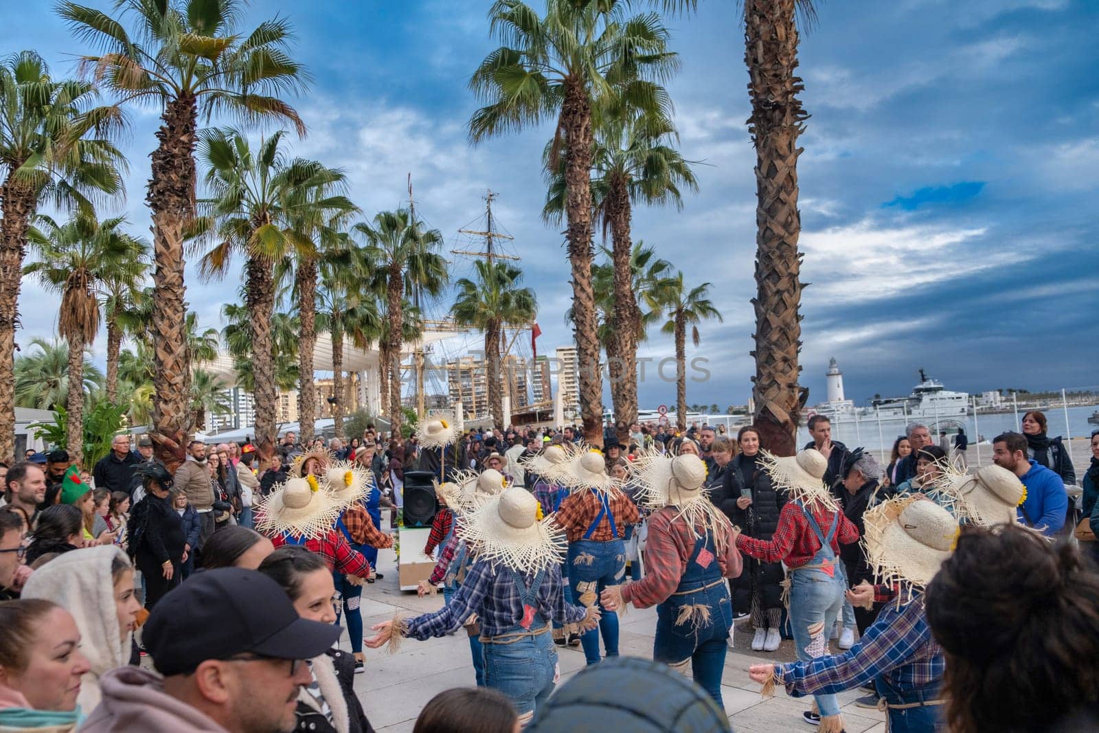 outdoor scene with a group of people engaged in what appears to be a dance performance or social dance gathering. They are wearing large straw hats, which are often associated with traditional or folkloric dance styles from various cultures. The participants are holding hands and appear to be performing a dance in a circular formation. Spectators surround the dancers, watching the performance