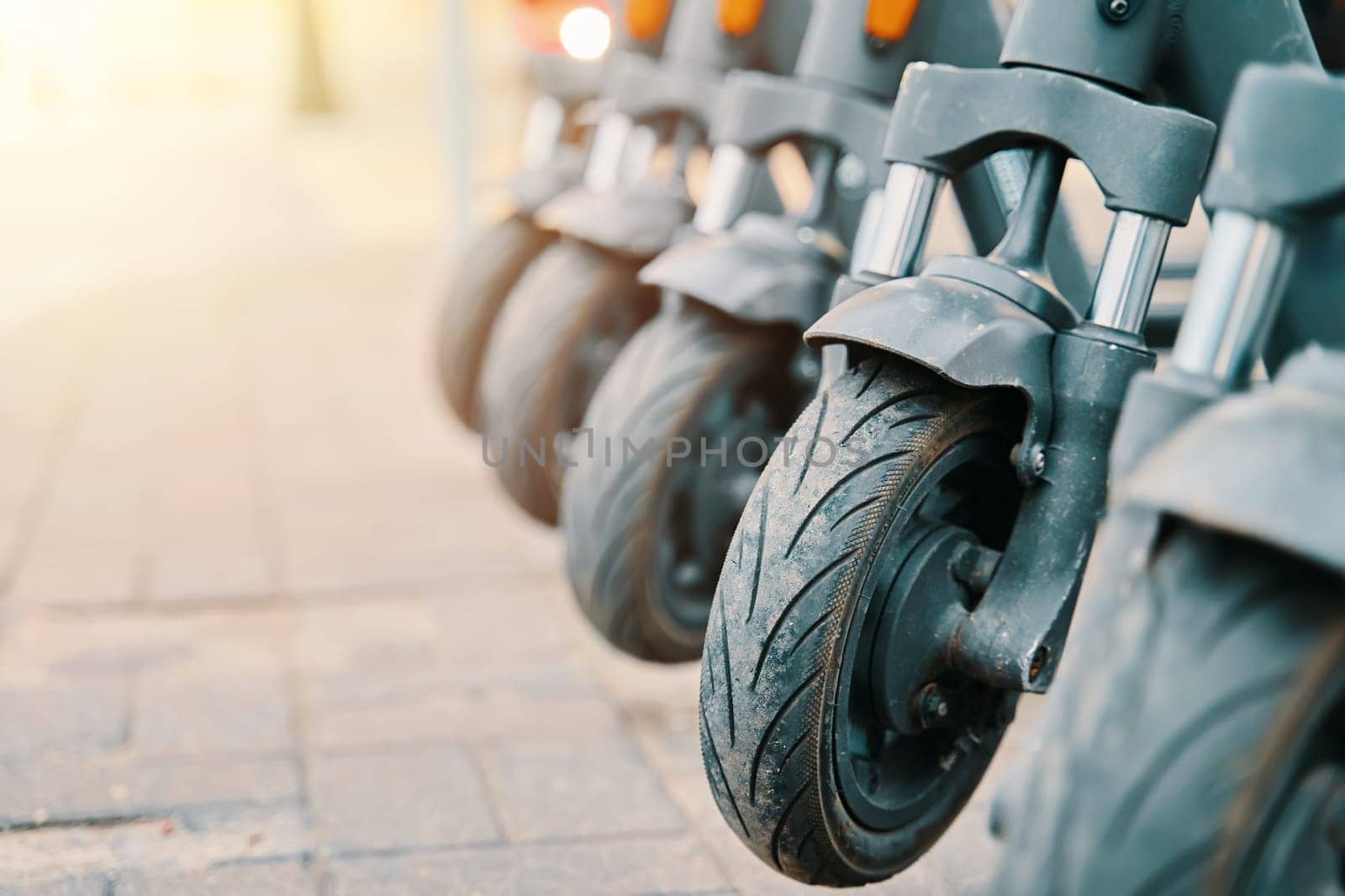 A line of scooters are parked on the sidewalk, resembling a sculptural row of art pieces made from metal, wood, and automotive tire materials