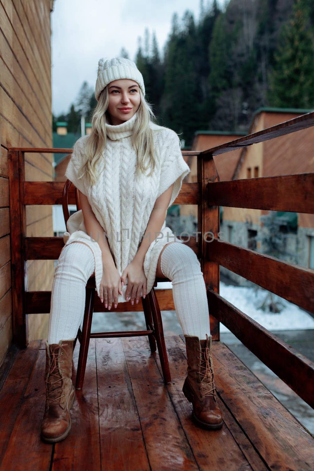 A sexy girl in a white sweater and stockings on the balcony admires the beauty of nature. wool clothes and a hat on a beautiful blonde.
