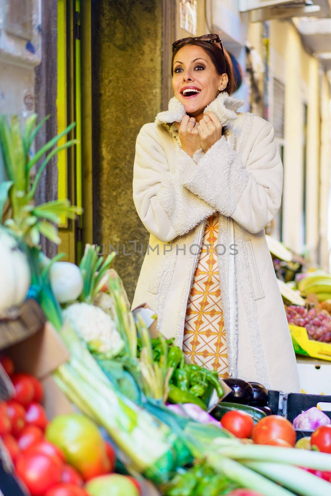 Positive young female greengrocer, in casual warm clothes with bunch of fresh fruits and vegetables looking at camera while standing and making punch fist in daylight