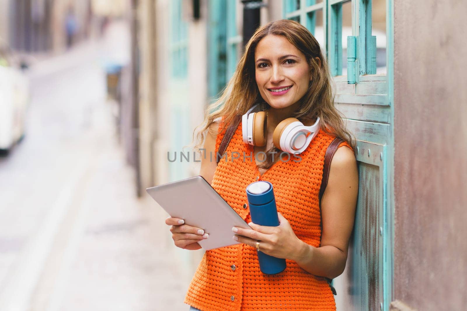 Latin happy woman holding reusable water bottle while using digital tablet in the city