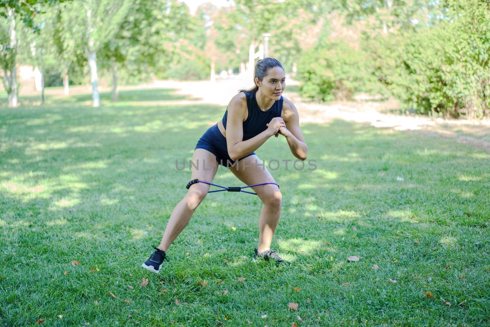 A young woman in sportswear performs exercises in a park with a resistance band, emphasizing fitness and wellbeing amidst greenery, focusing on maintaining her form and staying motivated