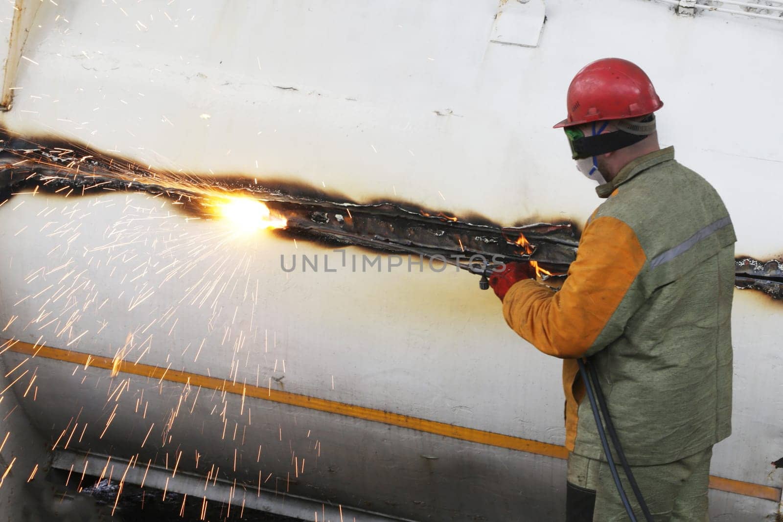 A male worker wearing overalls and a helmet with an autogen torch in his hands cuts a large metal item for disposal.