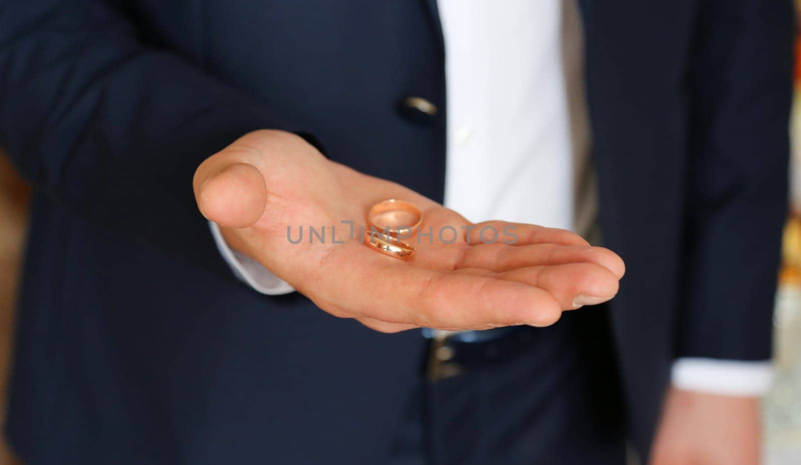 The groom in a blue suit and white shirt holds two gold wedding rings in the palm of his hand.