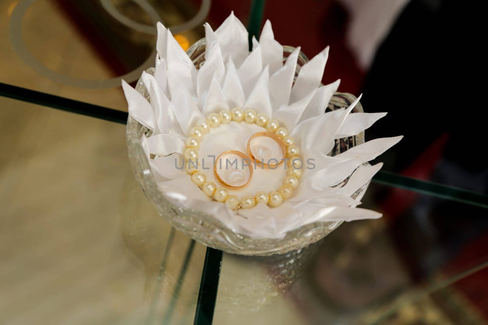 Two gold wedding rings and a pearl necklace lie on a transparent glass table, in a crystal dish covered with a decorative white napkin.