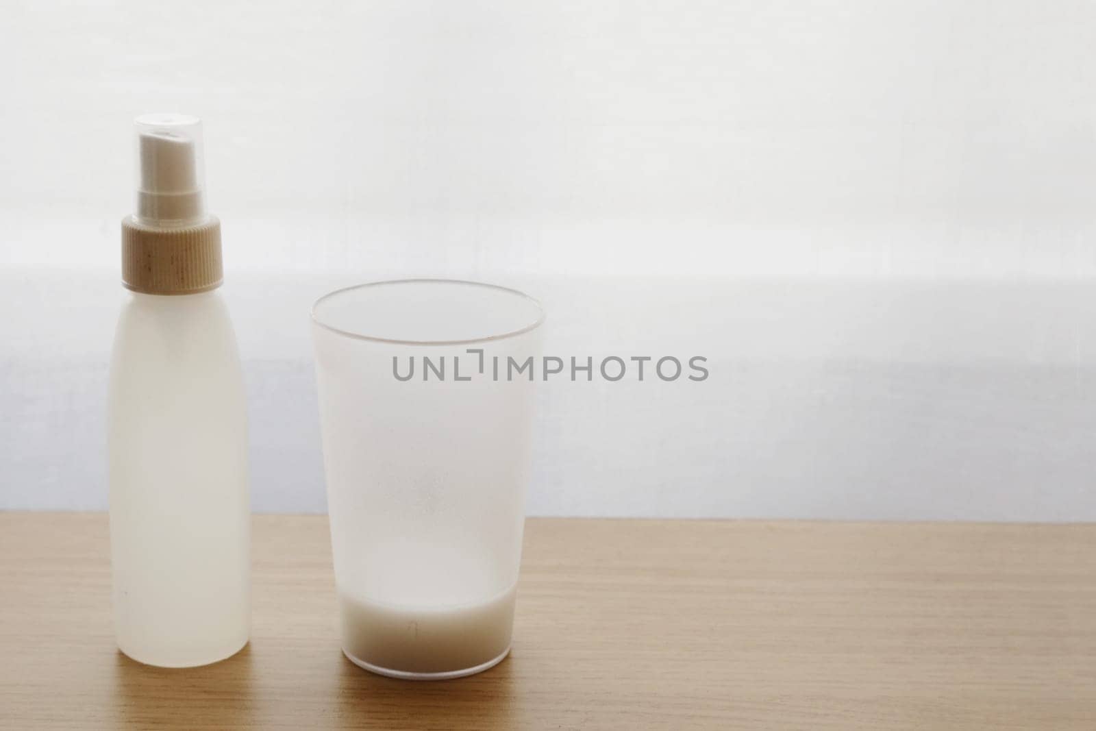Small sanitary bottle with antiseptic, plastic cup with liquid in the background of curtains and window.