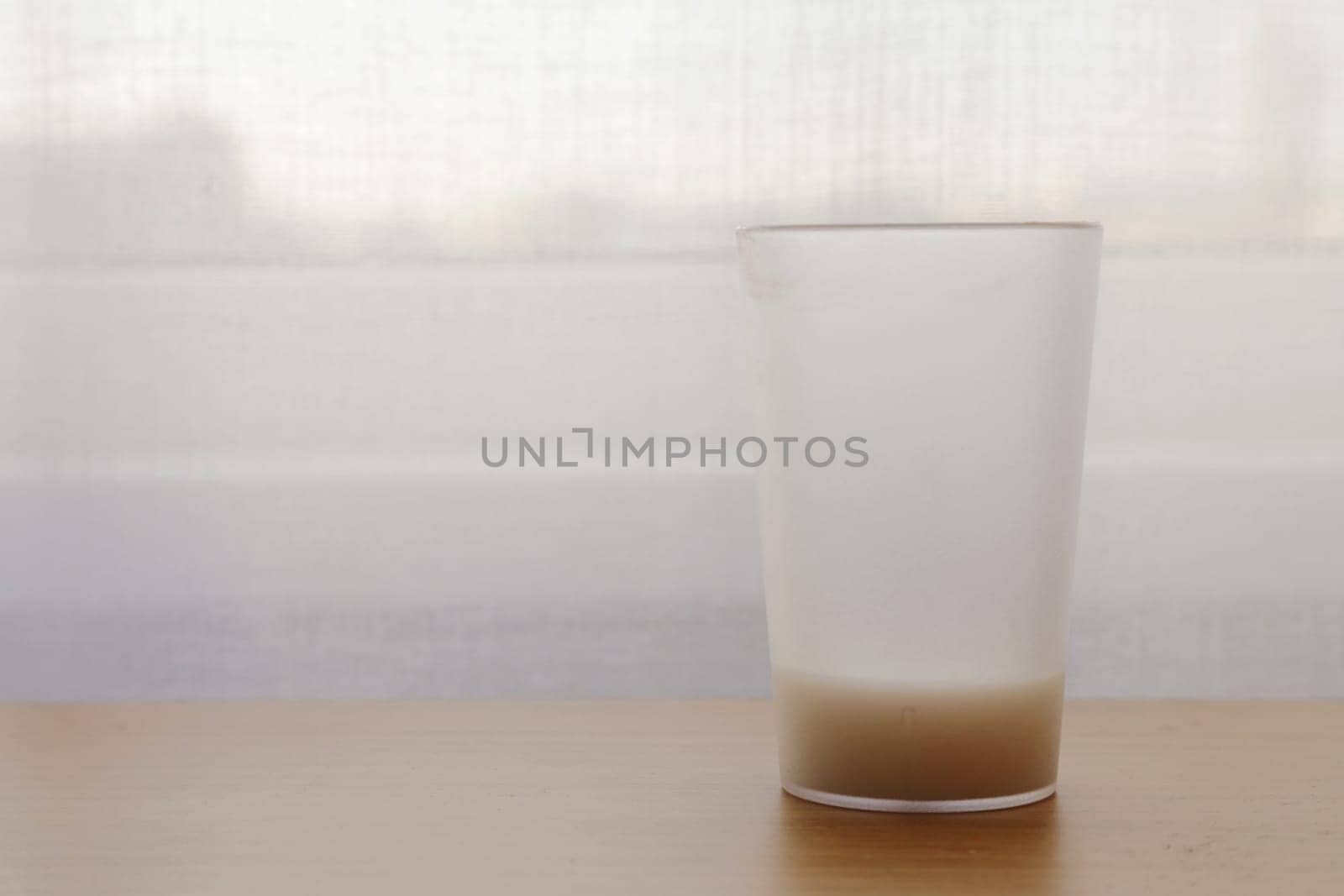 A large glass with an unfinished drink on the table against the background of a white window with a curtain.