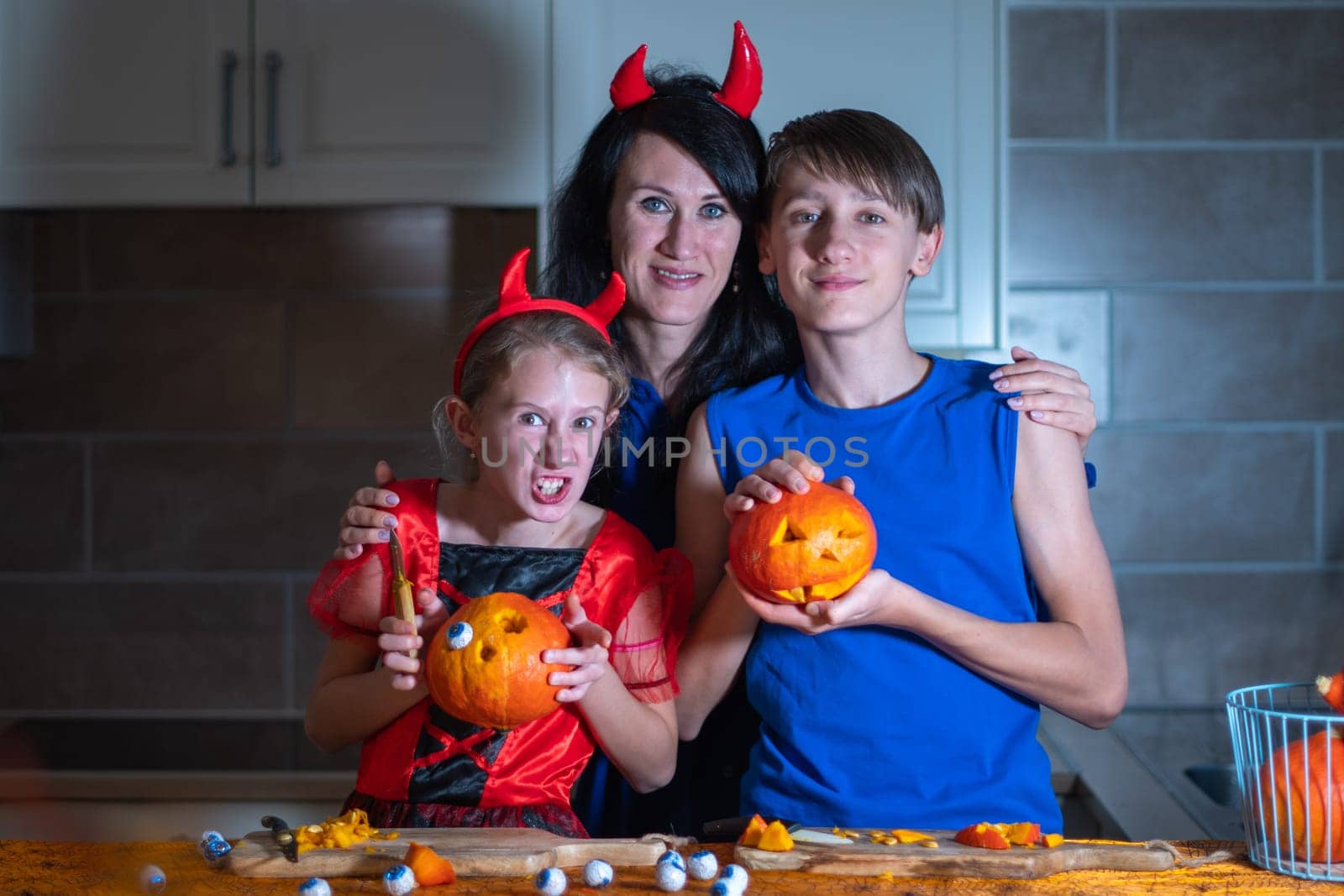 Mother, daughter and son in Halloween costumes carve eyes and teeth on pumpkins in a darkened room, traditional autumn decorations for the holiday