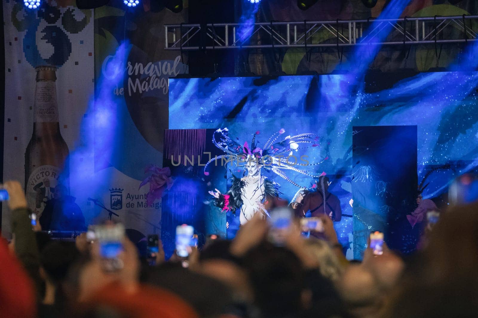 festive event or performance. In the forefront, there are many people holding up their phones, presumably taking pictures or recording the event. In the background, there's a brightly illuminated stage with blue hues and a figure that appears to be part of a performance or show