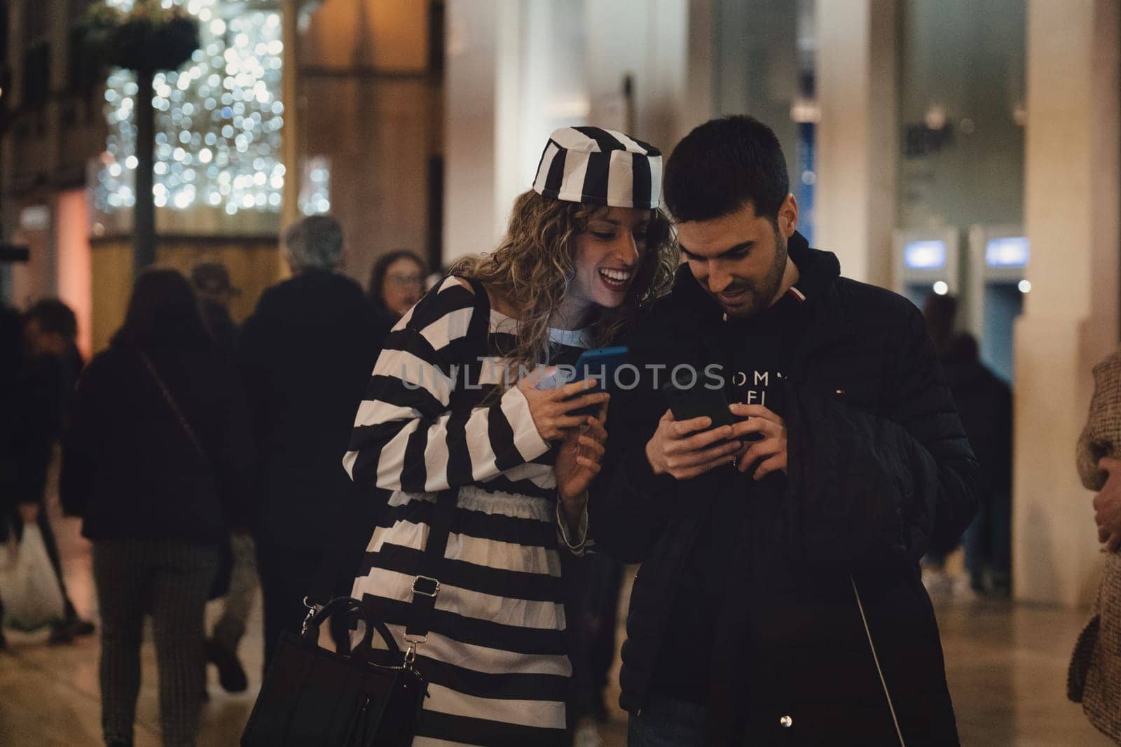 woman and a man standing close together looking at a smartphone, which the man is holding. The woman is smiling. She is wearing a striped garment that is black and white, and also a hat with similar stripes. The man is wearing a dark jacket. High quality photo