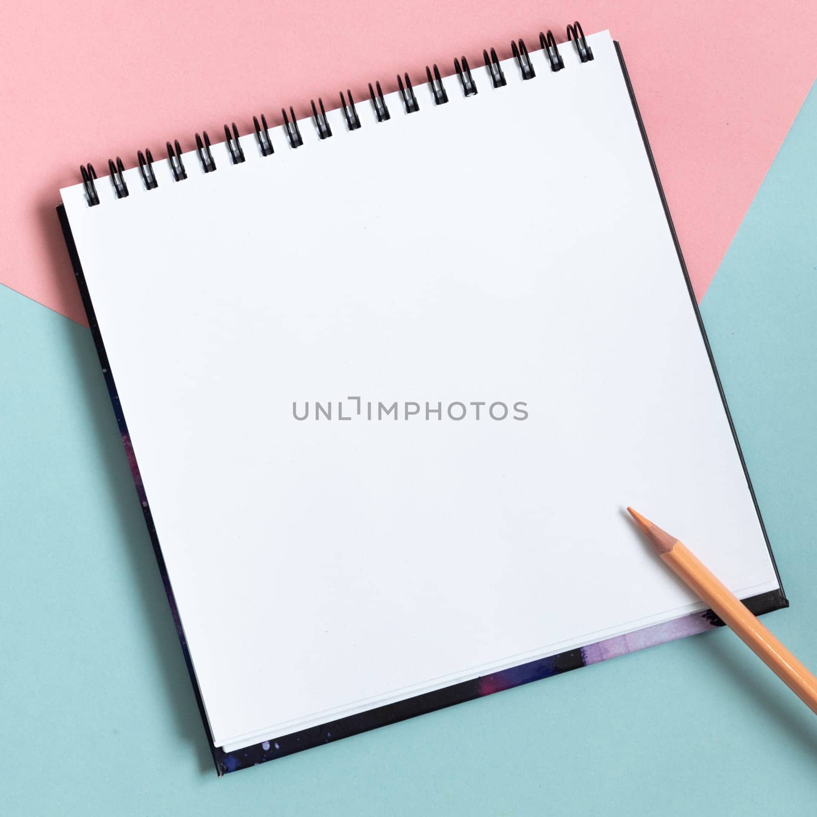 Mock-up of empty spiral sketchbook on pastel pink and blue background. Top view of open notebook with clean sheet and pencil. Template for message. Copy space