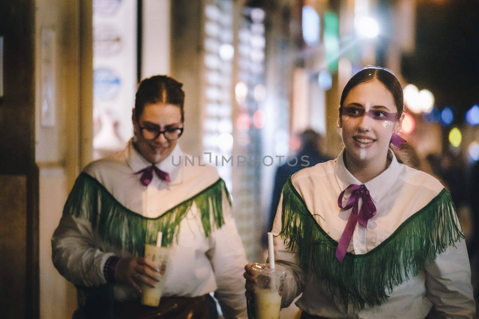 Women dressed in traditional or costume attire walking. both wearing white shirts with green fringe details on the shoulders and purple neckties. smiling and looking towards the camera. Each of them is holding a beverage