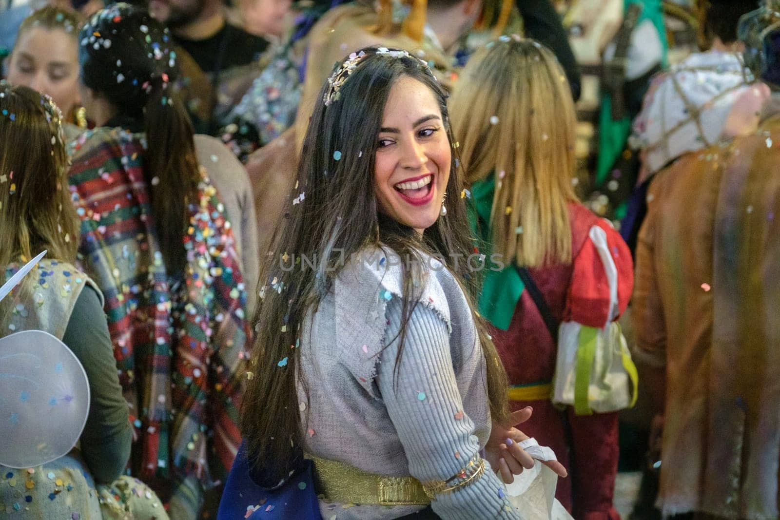 woman smiling broadly and looking over her shoulder, with what appears to be confetti scattered in her hair and on her clothes, she is at a festive event or celebration. Around her are several other people download image