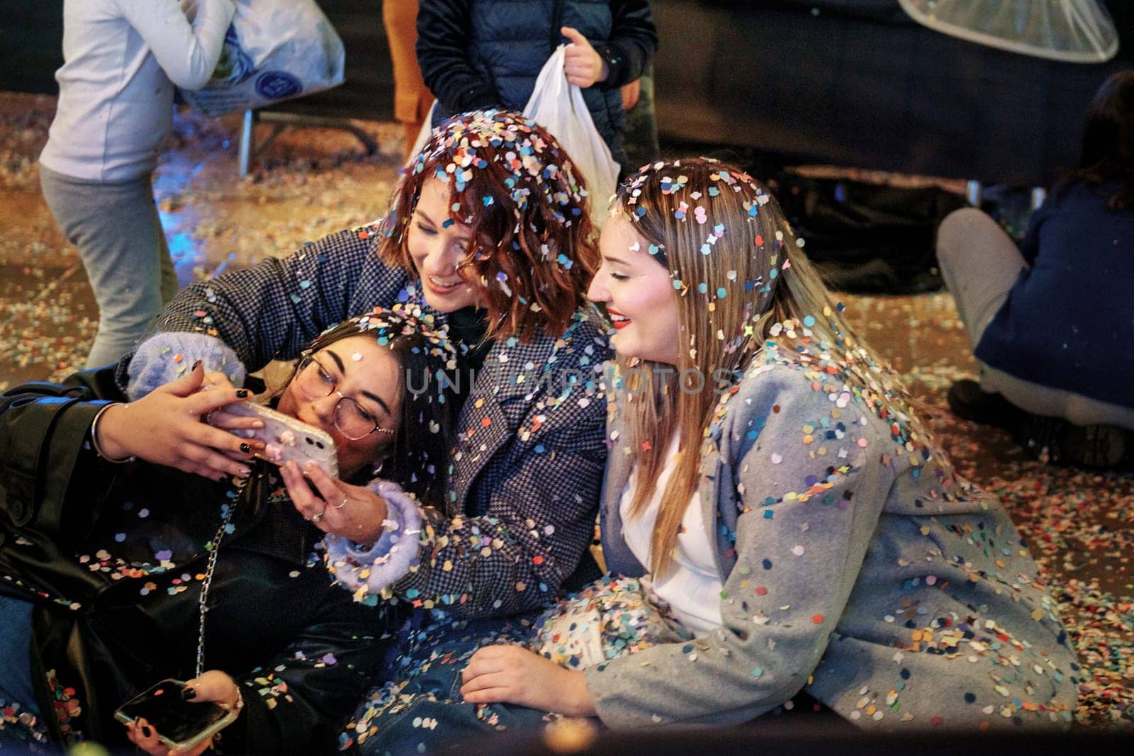 three woman taking a selfie together. They appear to be in a celebratory or festive environment, as they are covered with colorful confetti. The mood joyful and playful, as they are smiling and looking at the phone screen while capturing the moment