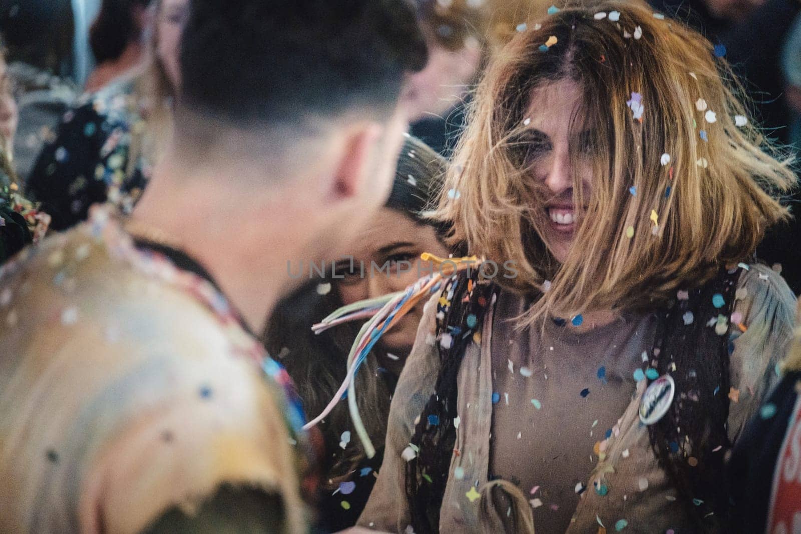 people in a festive setting, surrounded by confetti. They appear to be in the midst of a celebration, party or a festival. One person in the foreground is facing the camera with a joyful, lively expression and confetti is scattered on their hair and clothes