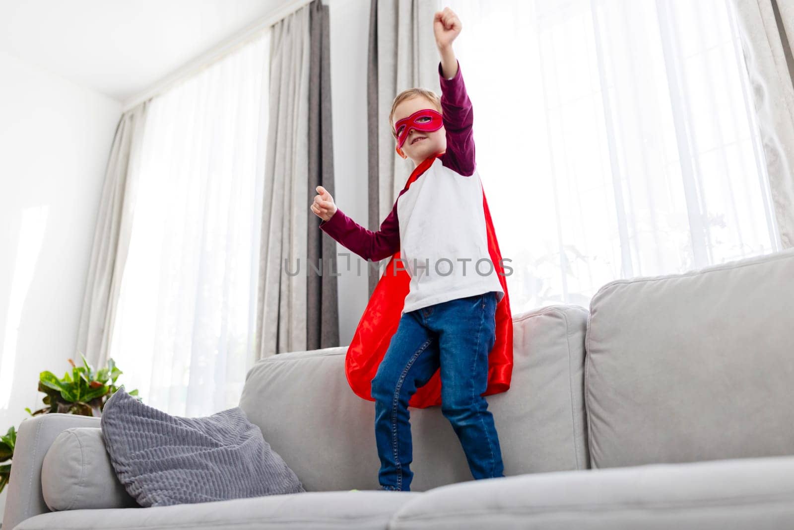 A boy dressed in a superhero costume with a red cape and mask standing on a grey couch. The child poses with arms raised triumphantly. The scene is set in a bright living room.