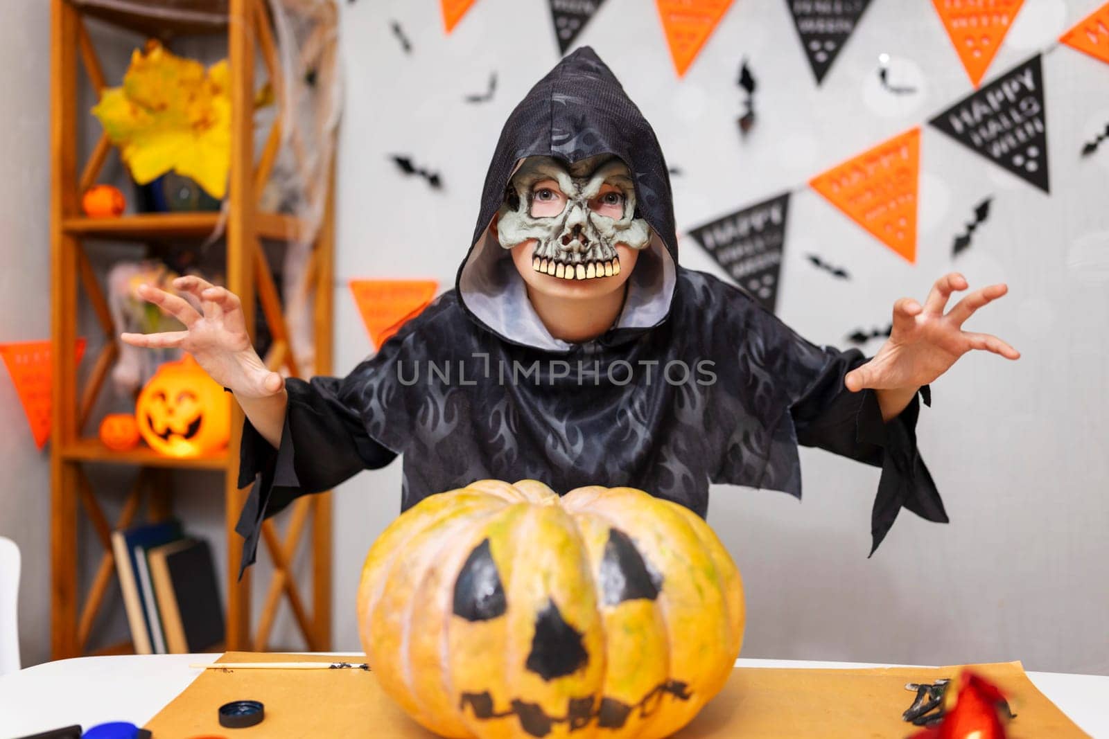 A child dressed in a skeleton costume poses menacingly with a carved pumpkin on the table. Halloween decorations, including banners and bats, adorn the background. Indoor setting.