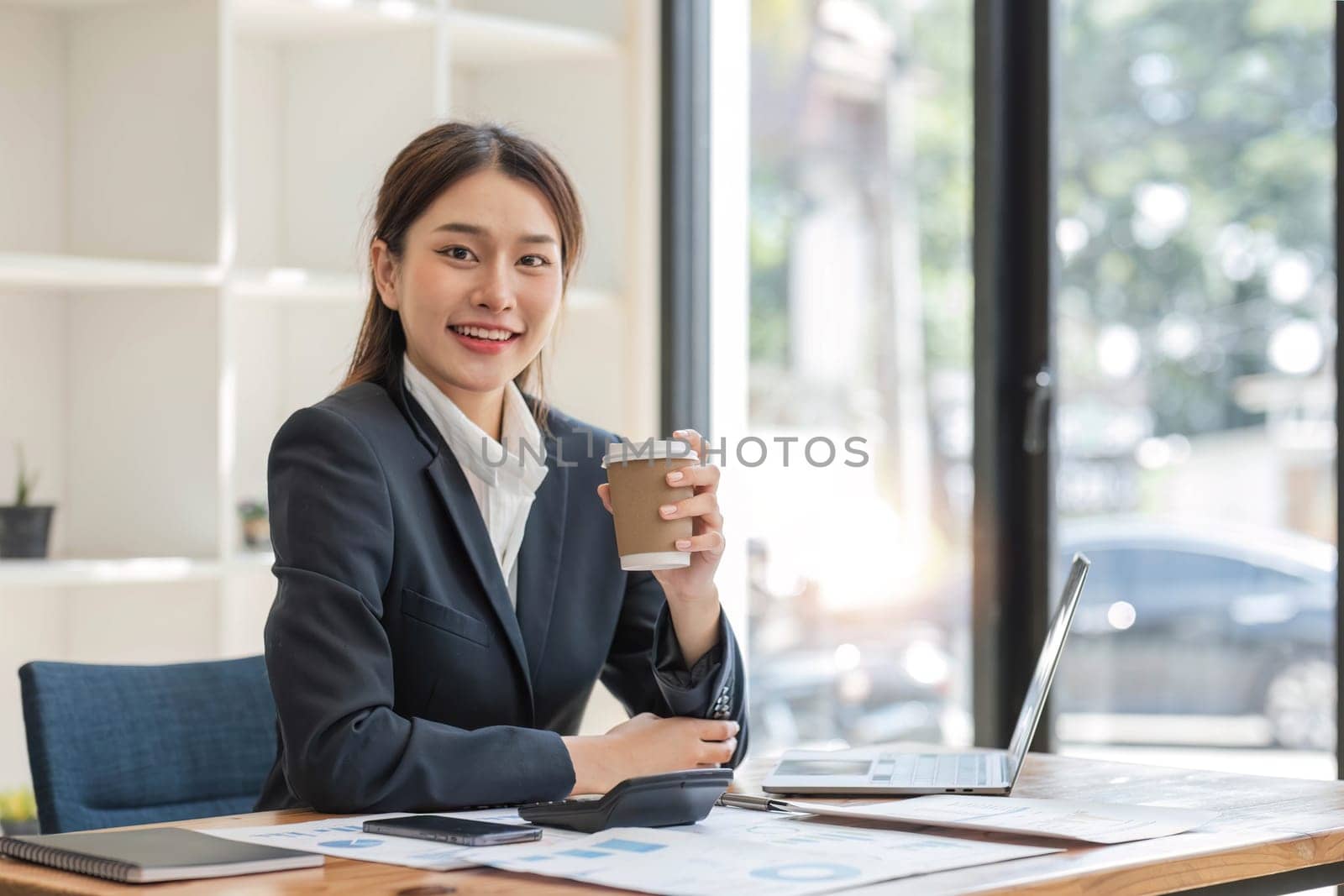 Photo of positive Asian businesswoman holds a cup of coffee imagines plan holiday in office. by wichayada