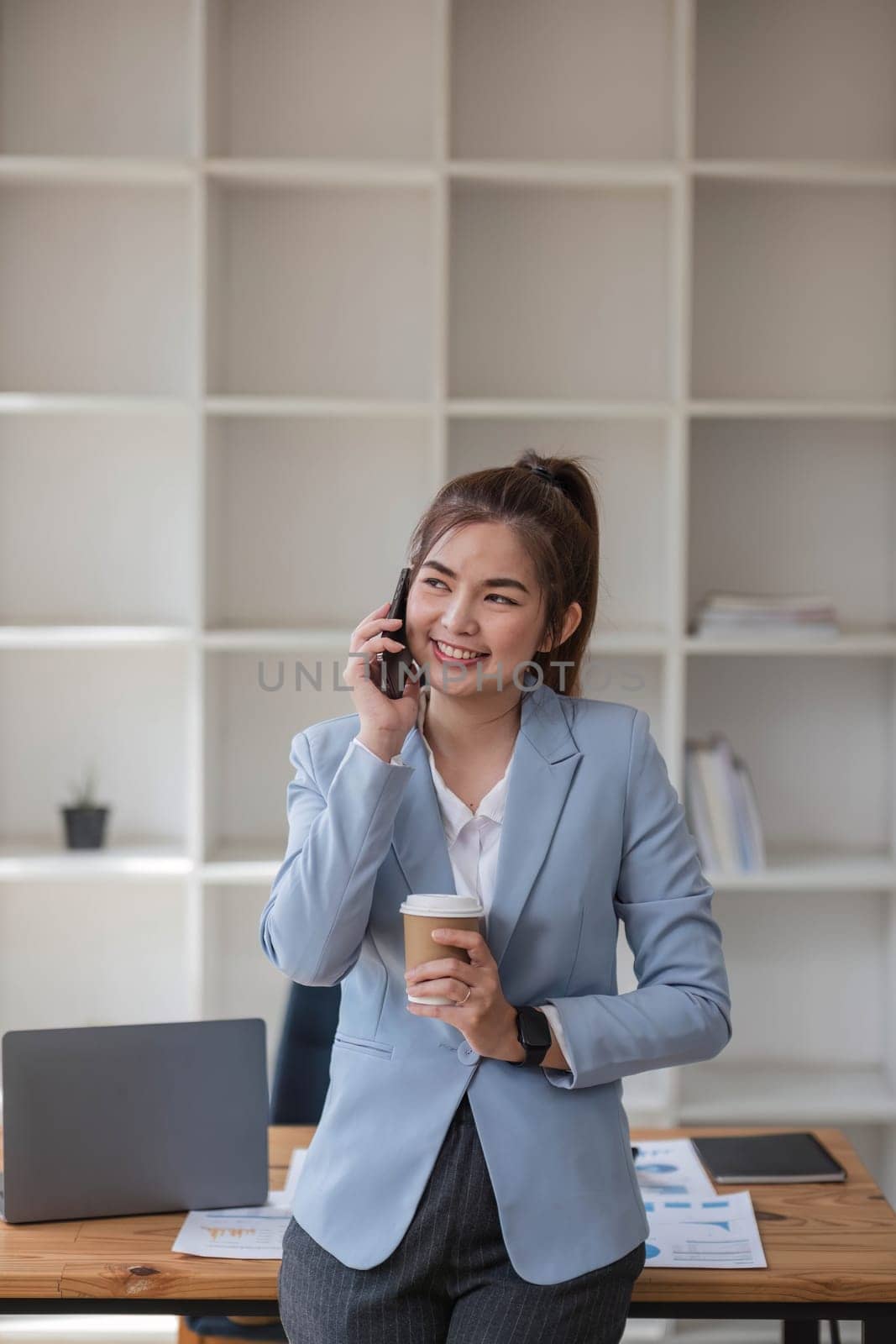 Asian businesswoman using mobile phone Smiling and happy while standing at desk in modern office by wichayada