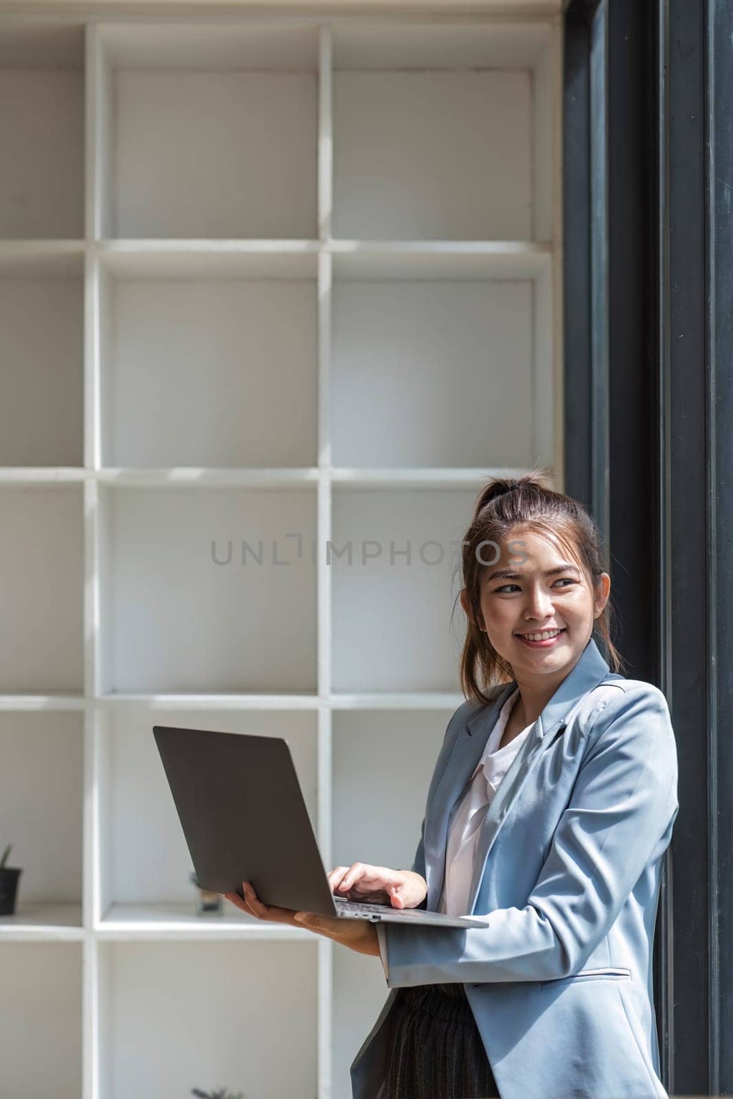 Portrait, Professional and confident Asian businesswoman or female executive manager in formal suit and use laptop. standing, leaning on table, holding laptop and using laptop computer. by wichayada