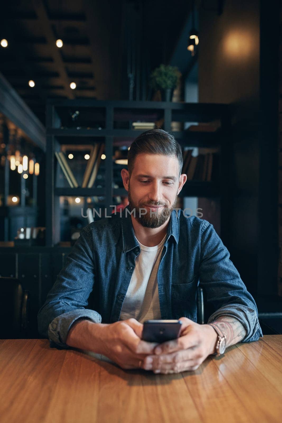 Young bearded businessman,dressed in a denim shirt, sitting at table in cafe and use smartphone. Man using gadget. by nazarovsergey