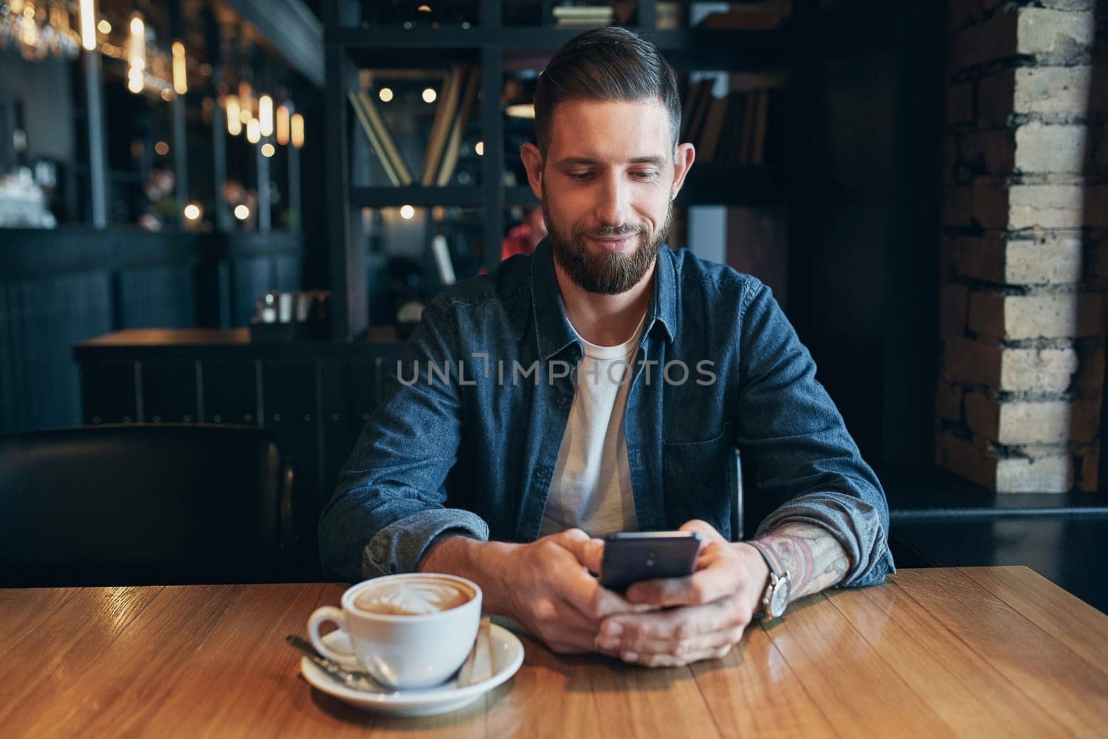 Young bearded businessman,dressed in a denim shirt, sitting at table in cafe and use smartphone. Man holding smartphone and looking at its screen. Man using gadget. Guy browsing internet on smartphone