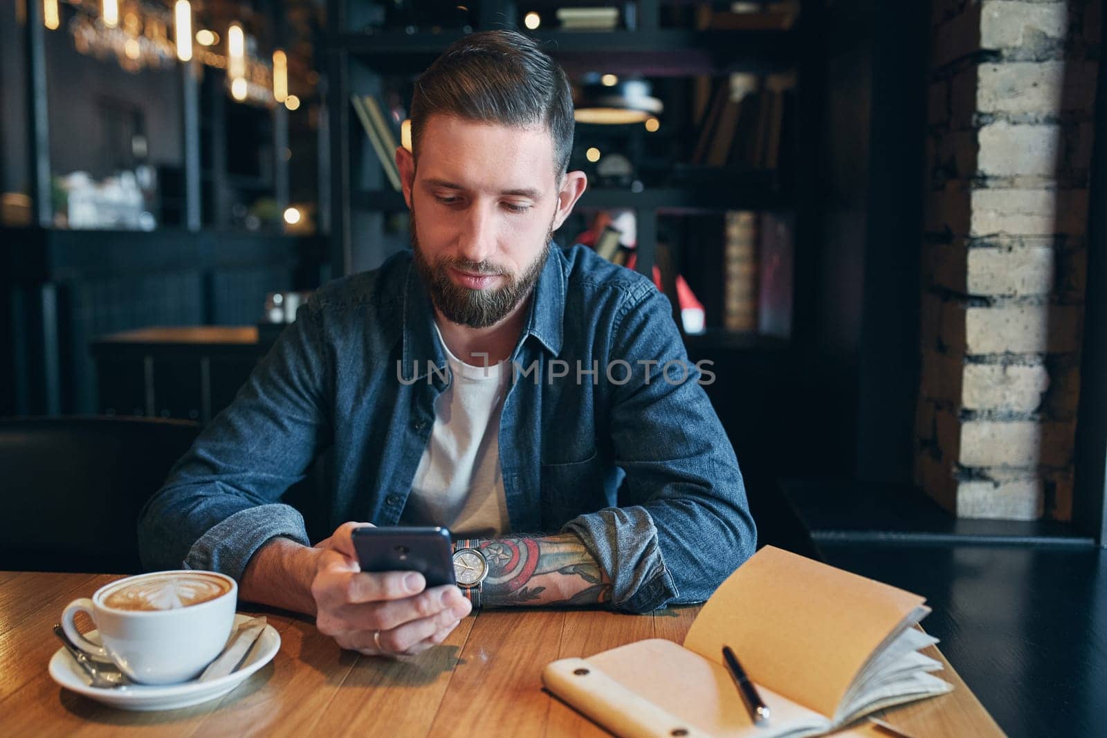 Young bearded businessman,dressed in a denim shirt, sitting at table in cafe and use smartphone. Man holding smartphone and looking at its screen. Man using gadget. Guy browsing internet on smartphone