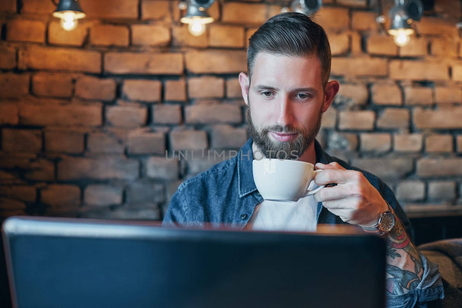 Young hipster guy at the bar having a cappuccino. Young man drinking coffee in city cafe during lunch time and working on laptop