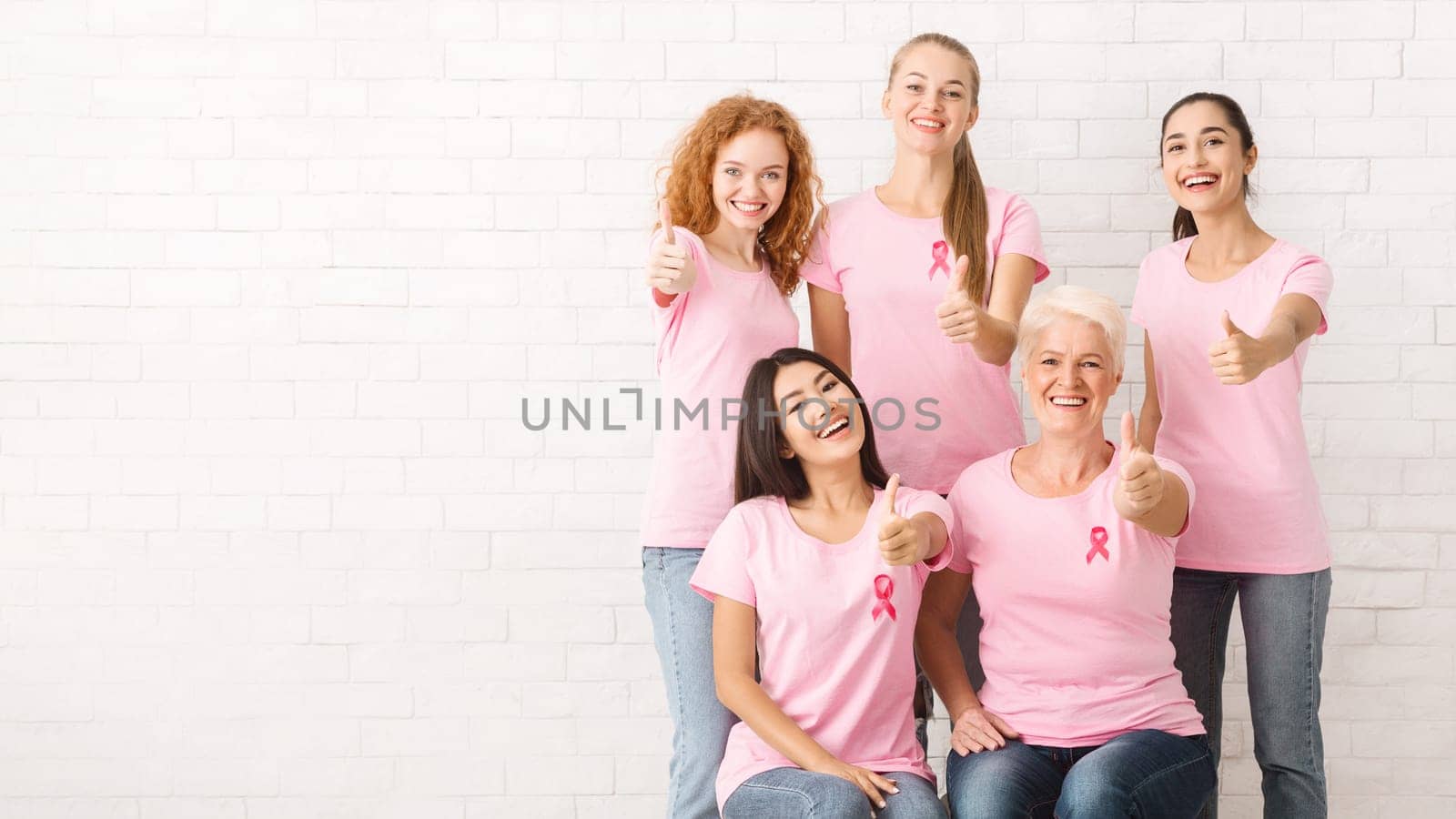 Diverse Female Volunteers In Pink Breast Cancer T-Shirts With Ribbons Gesturing Thumbs-Up Posing Over White Brick Wall. Copy Space