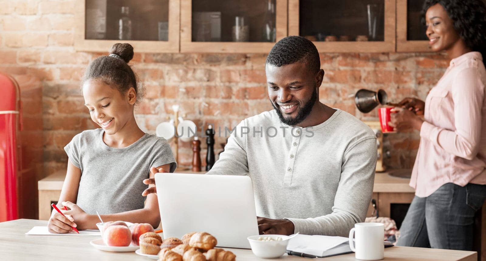 Happy afro family having breakfast together in kitchen, dad surfing internet on laptop, making plans for weekend, copy space