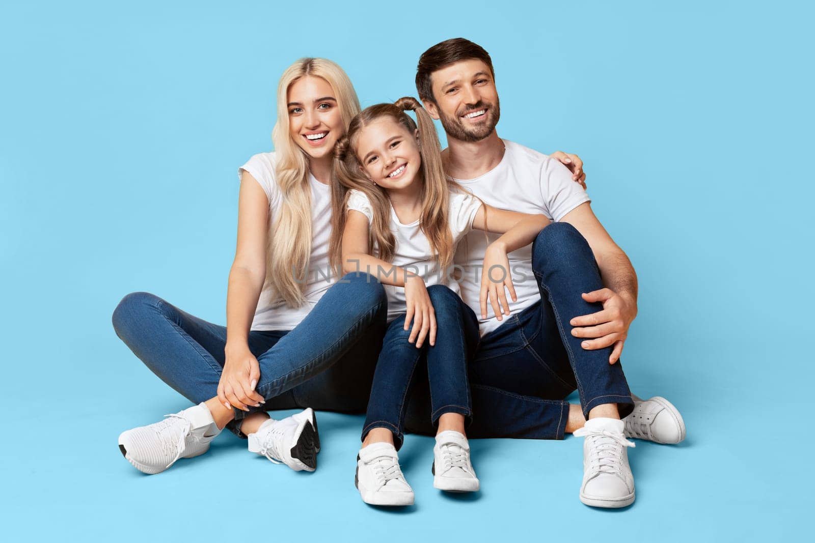 A young family, a mother, father, and daughter, are smiling and looking at the camera while sitting on a light blue background. They are wearing white T-shirts and jeans