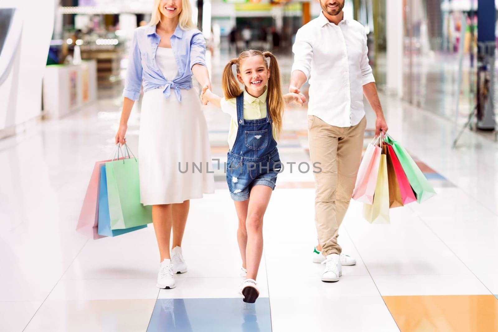 A family of three, a young girl with pigtails and her parents, are walking together through a modern shopping mall. The little girl holds onto both her mom and dad's hands