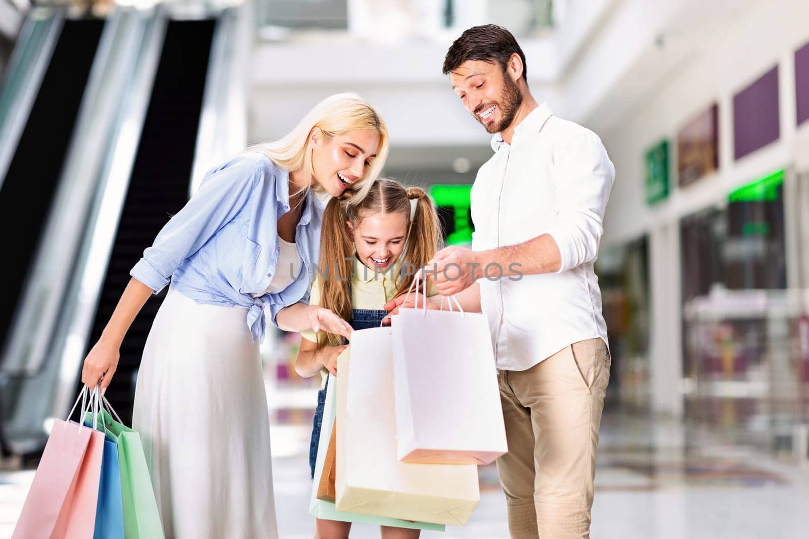 A young girl is standing between her parents, all three looking down at a pile of shopping bags. The girl is smiling, and the parents are smiling as they look at the contents of the bags.