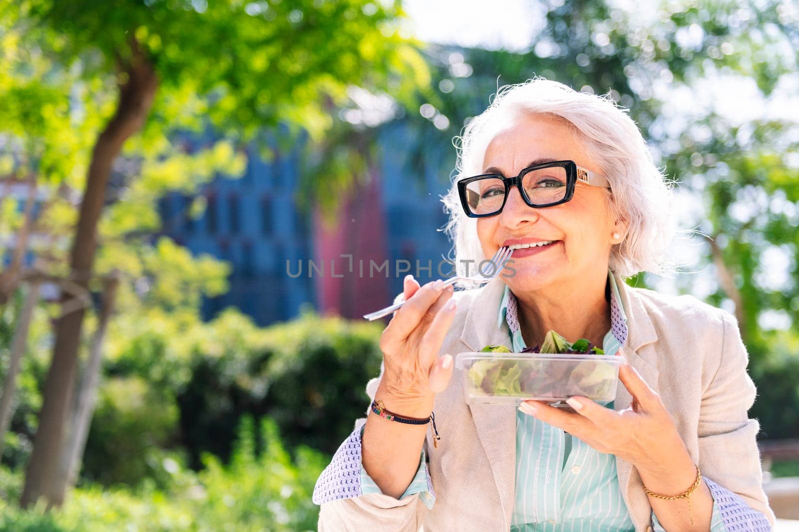 portrait of a middle age woman smiling happy while eating a takeaway salad sitting outdoors in a city park, concept of healthy food and active lifestyle in adulthood, copyspace for text