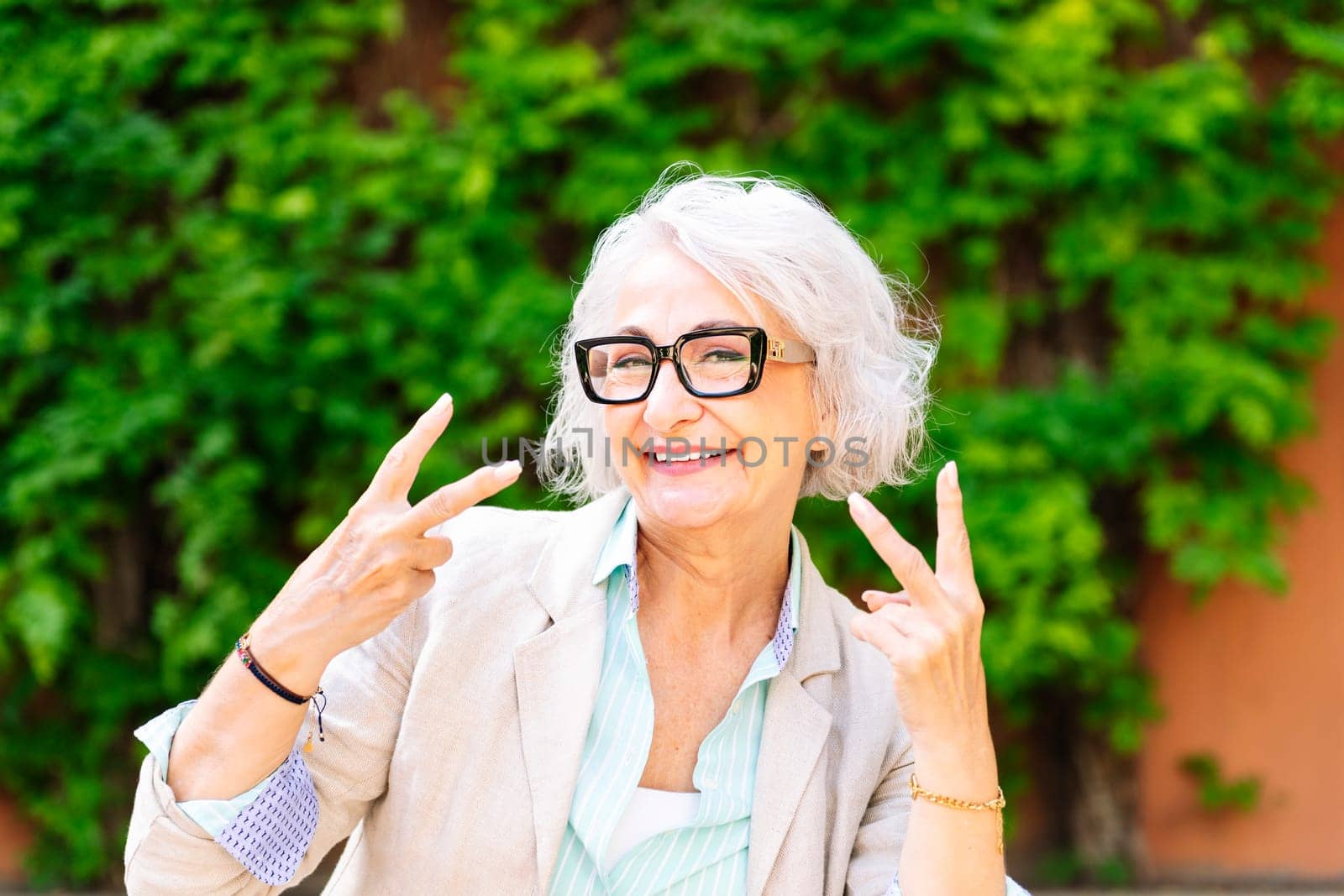 portrait of a middle age woman smiling happy and doing victory sign with hands while looking at camera with blurred green plants background, concept of mature people lifestyle