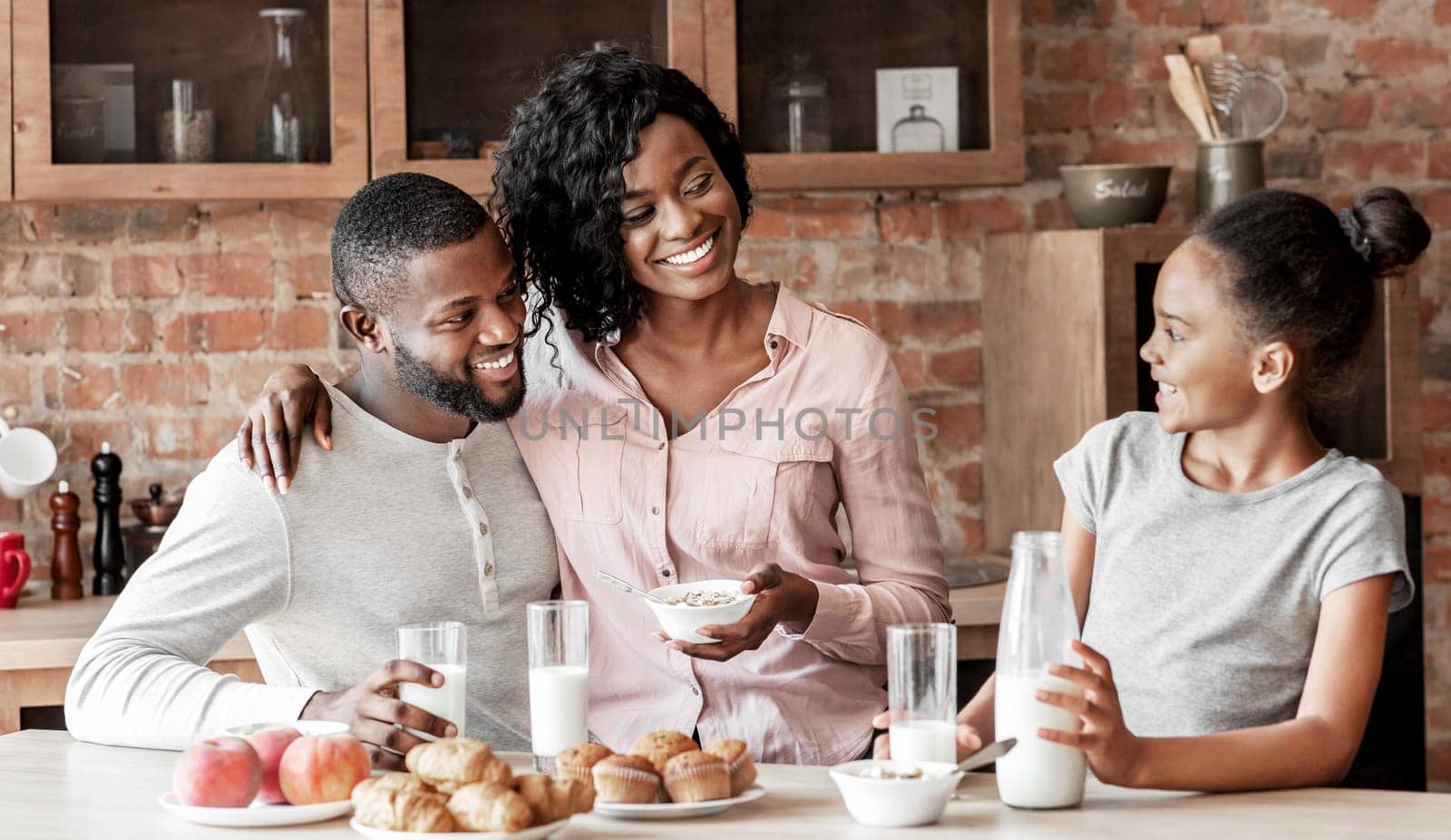 Adorable african family of three having breakfast at kitchen, eating and talking, copy space