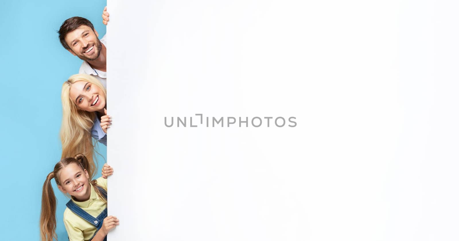 A family of three smiles brightly as they peek from behind a large white wall. The father stands at the top, followed by the mother, and their young daughter.