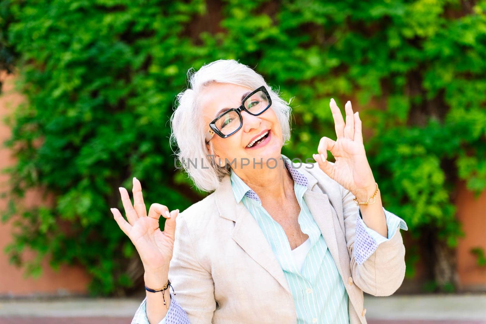 portrait of a middle age woman smiling happy and doing ok sign with hands while looking at camera with blurred green plants background, concept of mature people lifestyle