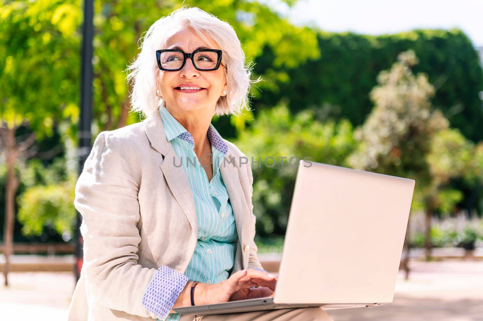 middle age woman smiling happy using laptop sitting outdoors in a city park, concept of technology and modern mature people lifestyle