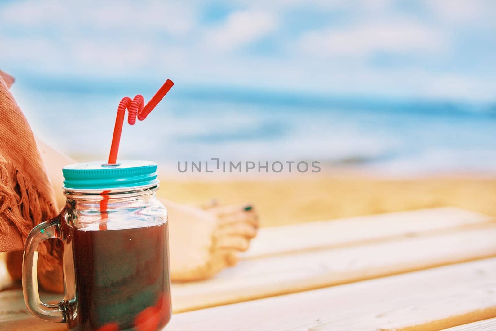 Glass jar with a drink and a red straw on a wooden chaise longue. Female feet in the background. Vacation on the tropical coast. Photo card with a copy space.