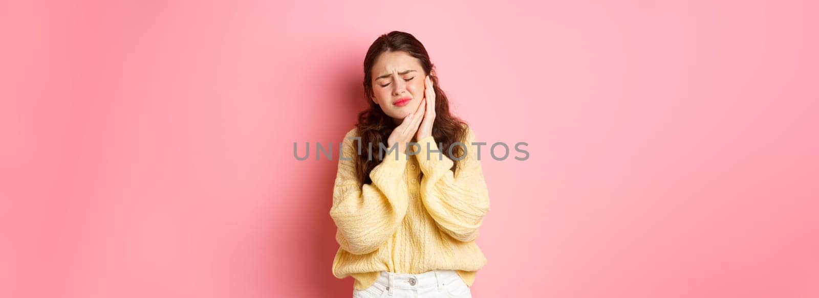 Young woman with terrible toothache, touching swallen cheek and grimacing from painful feeling, having tooth decay, need appointment to dentist, standing against pink background.