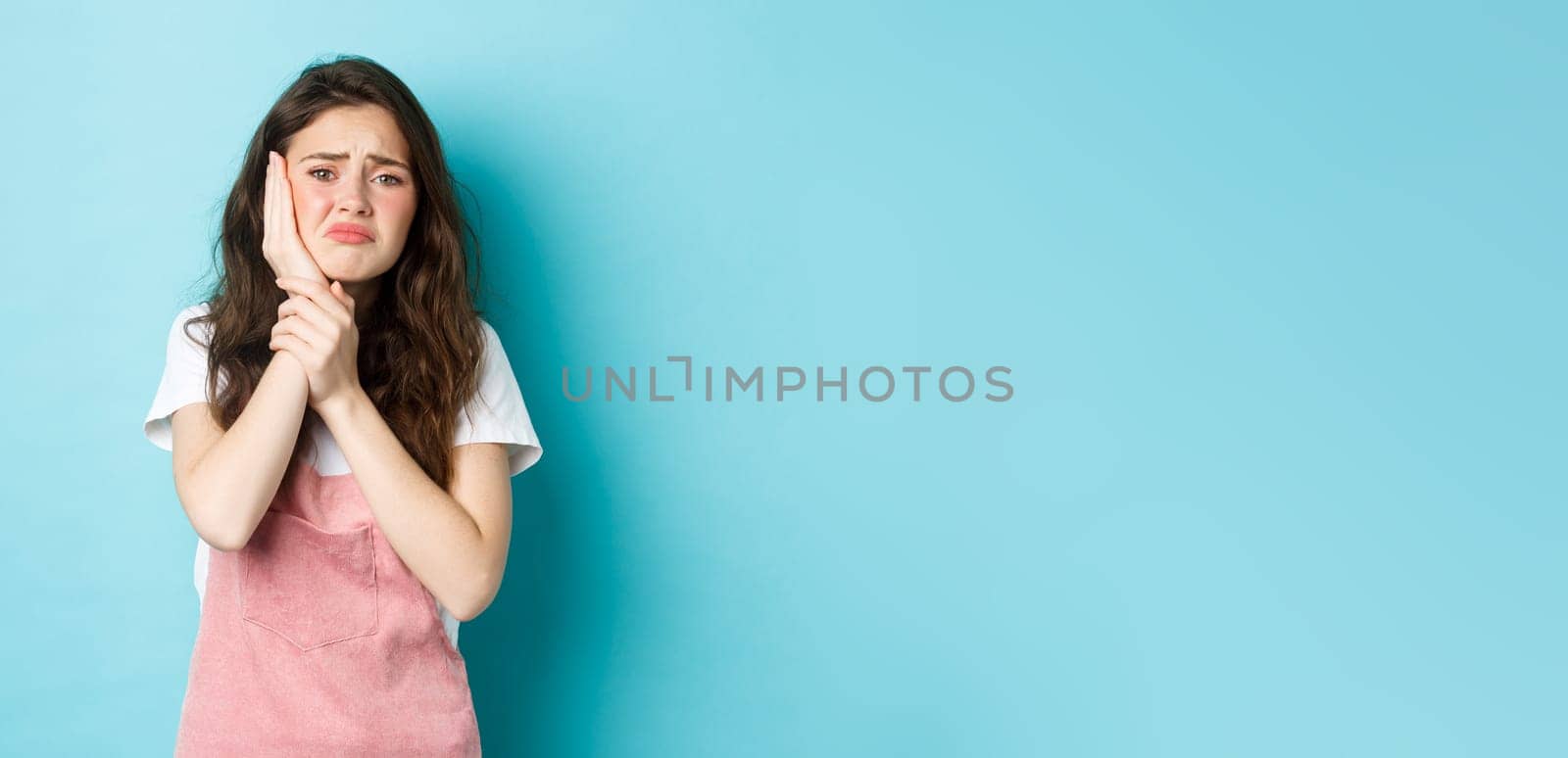 Image of frowning upset girl touching swallen cheek, having toothache, complaining on tooth decay, need to see dentist, standing against blue background.